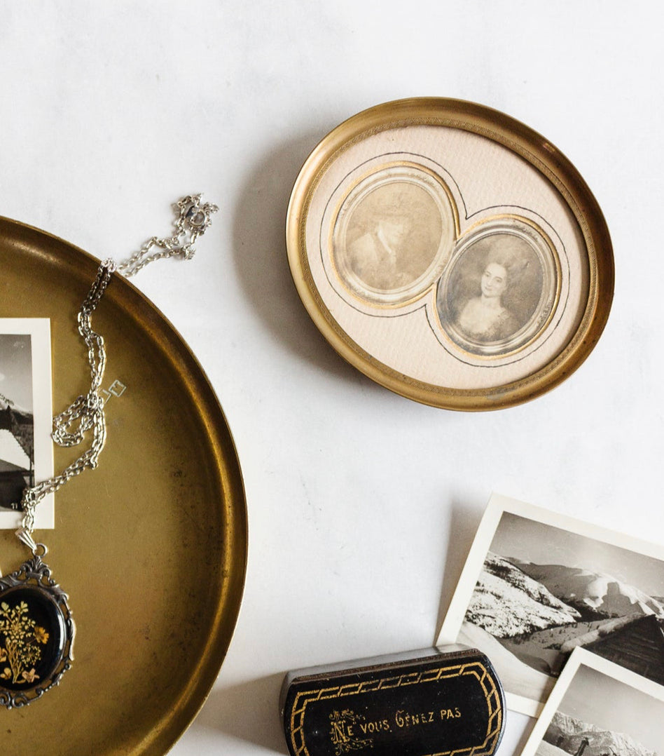 A 1920s French petite double portrait frame with French matting displays sepia portraits, surrounded by old black and white photos, a round gold tray, a silver locket necklace, and an antique black case on a white surface.