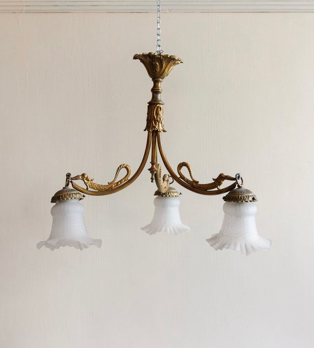 A 1920s French brass chinoiserie 3 light chandelier with ornate details and three frosted glass shades hangs from a chain against a plain, light-colored wall.