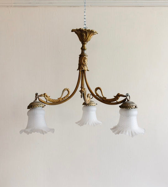 A 1920s French brass chinoiserie 3 light chandelier with ornate details and three frosted glass shades hangs from a chain against a plain, light-colored wall.