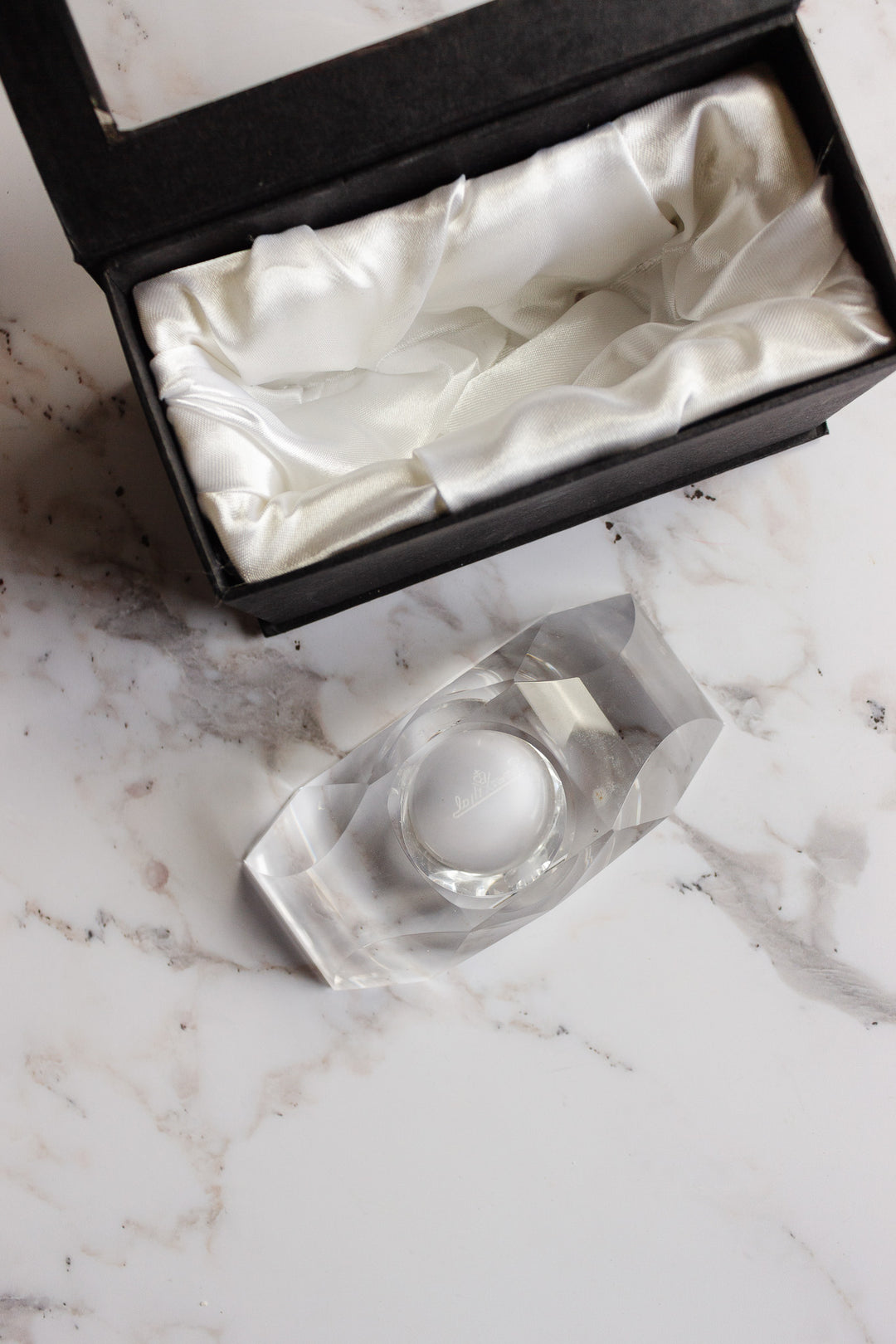 A vintage Rosenthal crystal votive rests on a marble surface beside an open black gift box lined with white satin.