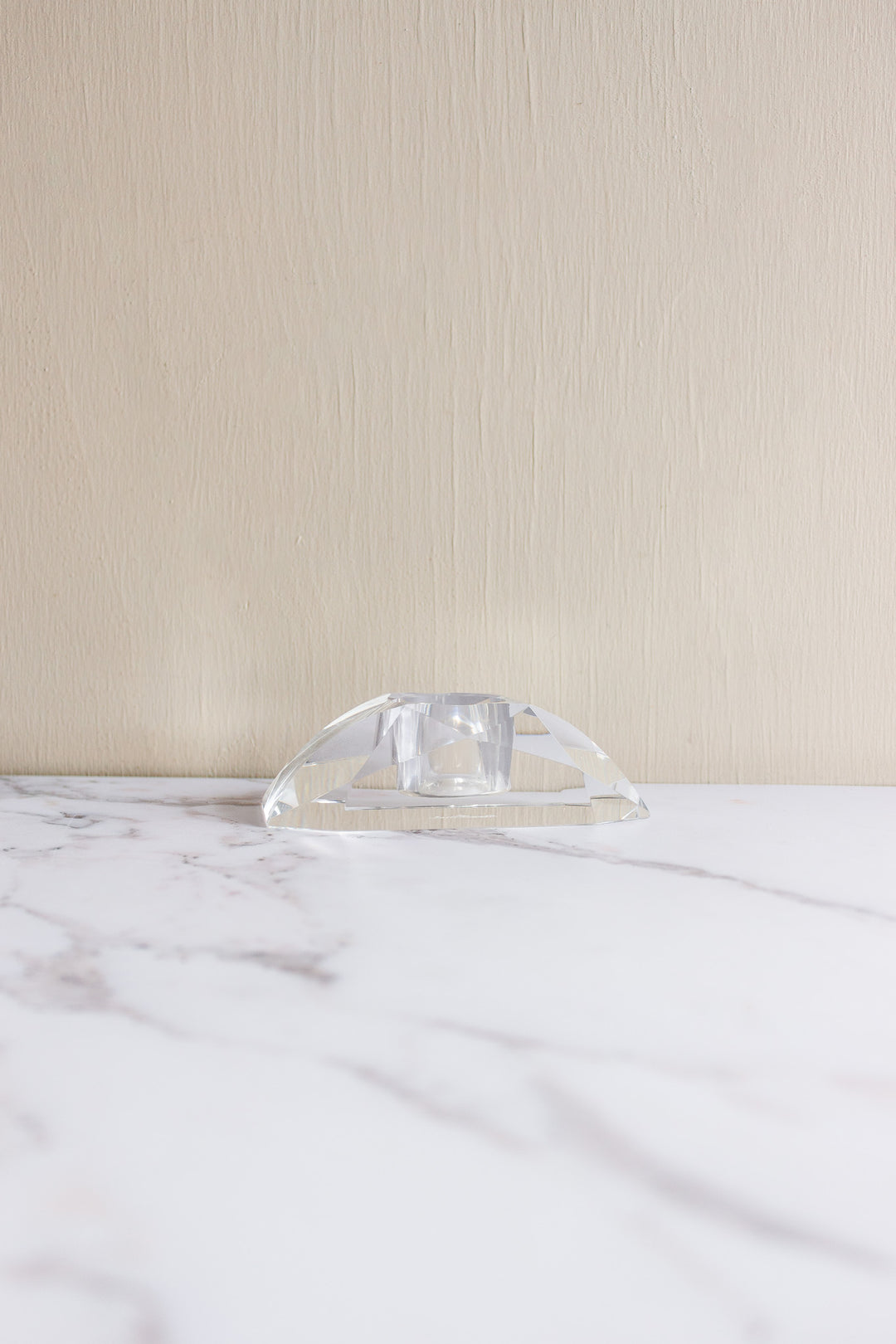 A vintage Rosenthal crystal votive, small and transparent with geometric facets, rests on a white marble surface against a beige textured wall, evoking the brand's refined elegance.