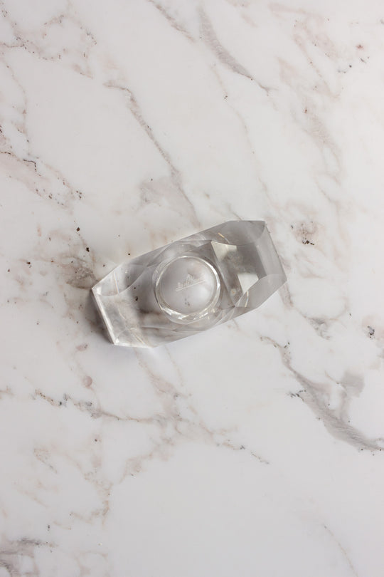 A clear tape dispenser with a roll of transparent tape sits on a white marble surface with gray veins, beside a vintage Rosenthal crystal votive.