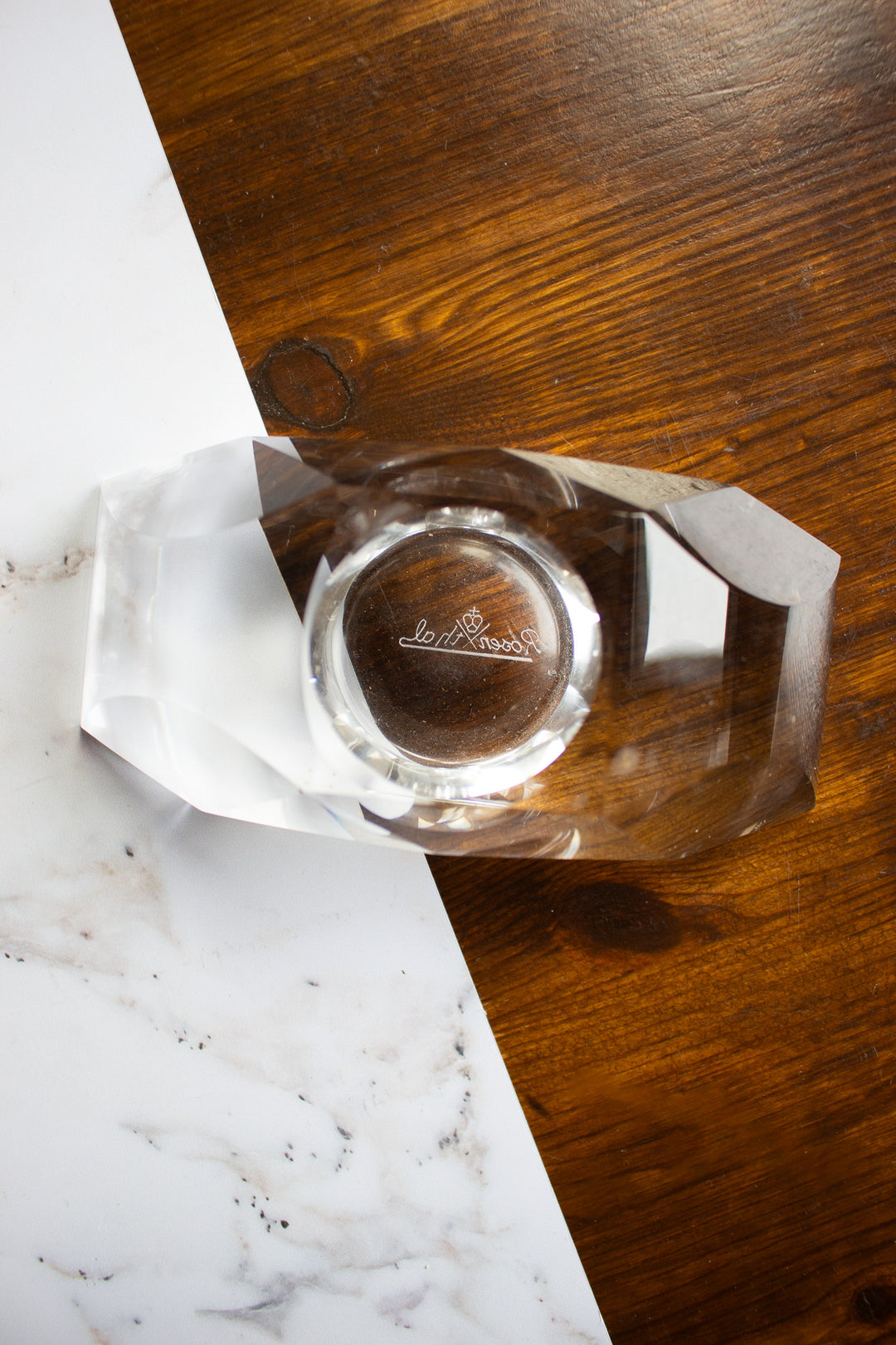 A vintage Rosenthal crystal votive featuring a faceted clear design with a brown circular center engraved with a motif rests on a surface that combines wood and white marble, reflecting the brand’s signature elegance.