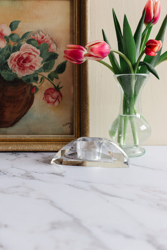 A clear glass vase with pink tulips sits on a marble surface beside a framed floral painting. In front of the vase, a vintage Rosenthal crystal votive adds sparkle and complements the elegant decor.