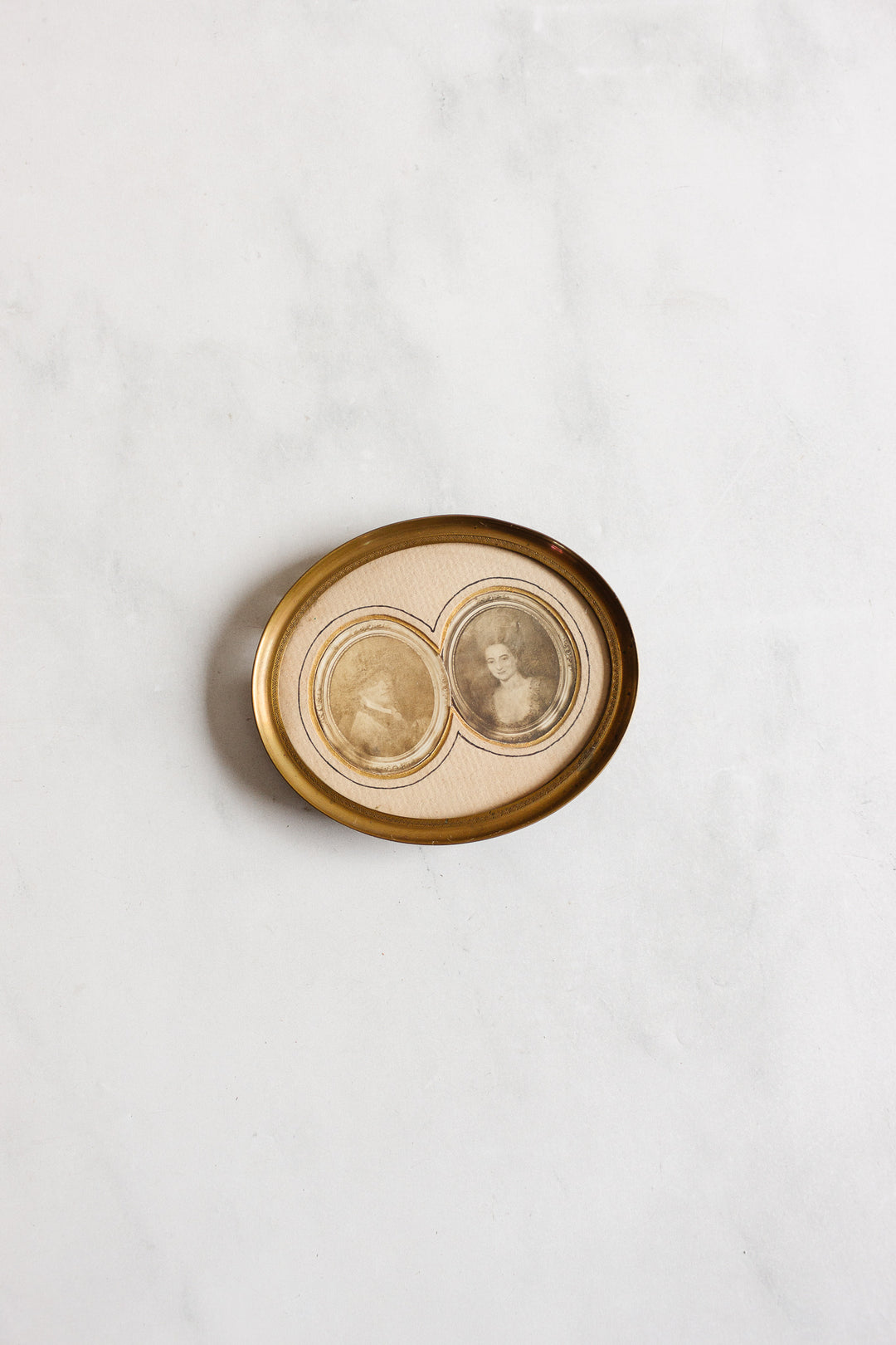 A 1920s French petite double portrait frame with French matting displays two sepia-toned vintage portrait photos side by side on a textured surface, creating a connected image within the antique brass frame.