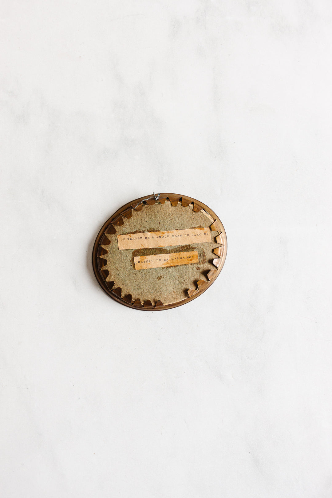 A 1920s French petite double portrait frame with French matting, oval-shaped with fabric backing and brown zigzag trim, displays two narrow strips of faded typewritten text and hangs on a white wall.