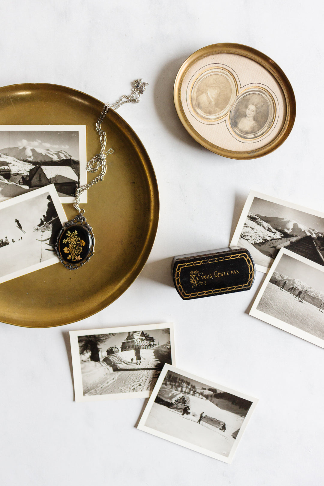 A set of vintage black-and-white photos on a white surface, featuring a gold tray with an ornate locket, a 1920s French petite double portrait frame with French matting, and a black and gold eyeglass case.