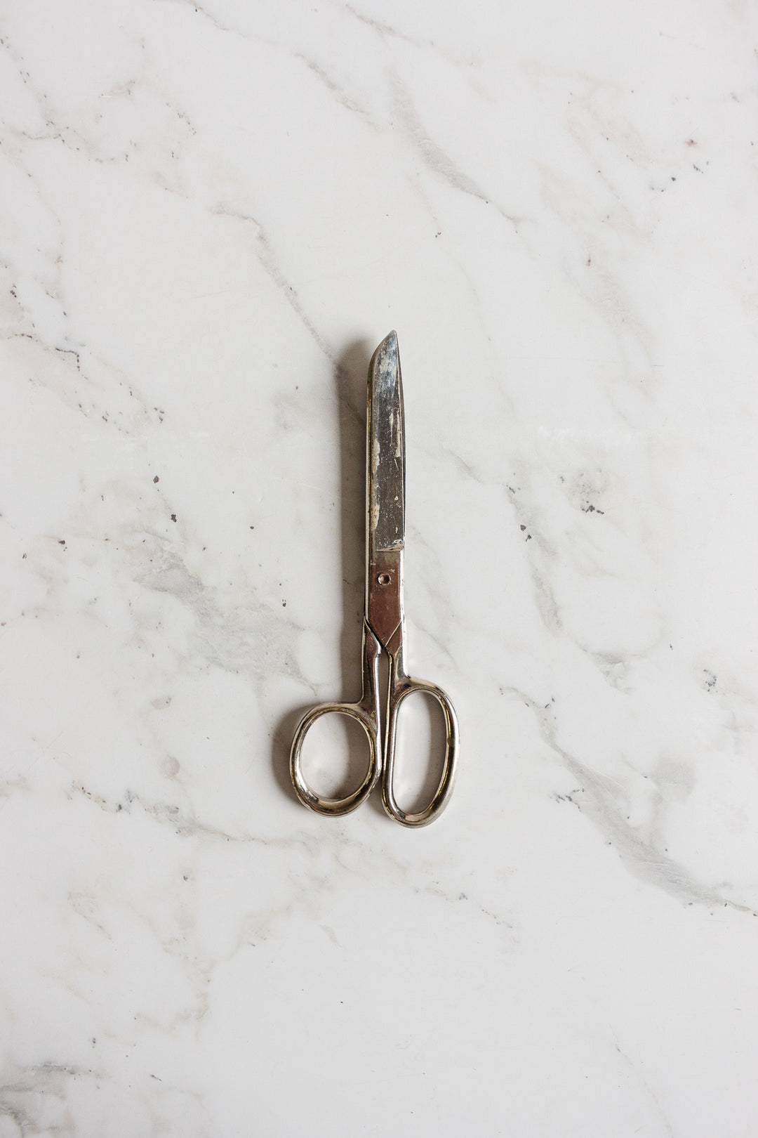 A pair of midcentury German scissors with a slightly tarnished finish rests closed on a white marble surface.