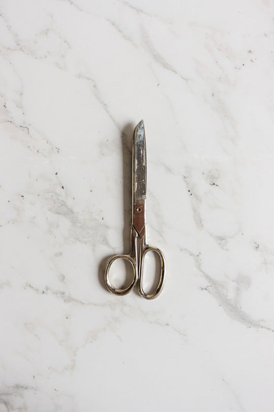 A pair of midcentury German scissors with a slightly tarnished finish rests closed on a white marble surface.