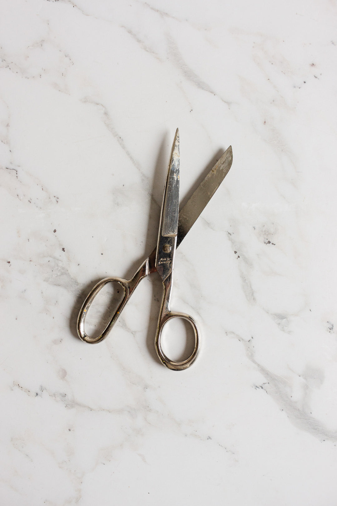 A pair of midcentury German scissors with slightly open blades lies on a white marble surface with light grey veining.