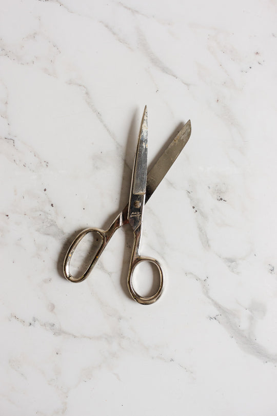 A pair of midcentury German scissors with slightly open blades lies on a white marble surface with light grey veining.