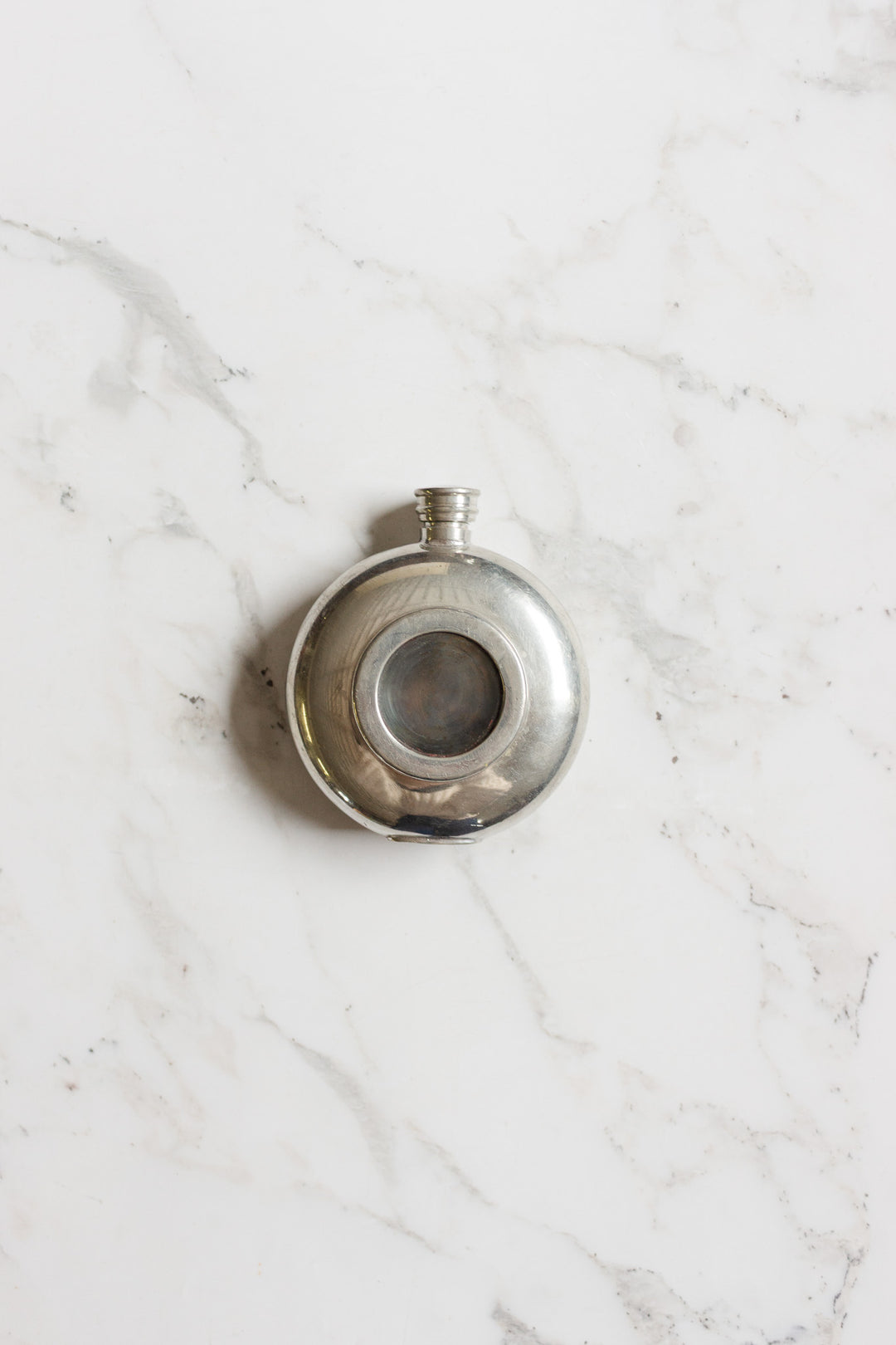 The flask is a round, silver metal container with a small screw cap and a circular glass window at its center, highlighting its compact size, displayed on a white marble surface.