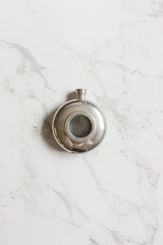 The flask is a round, silver metal container with a small screw cap and a circular glass window at its center, highlighting its compact size, displayed on a white marble surface.