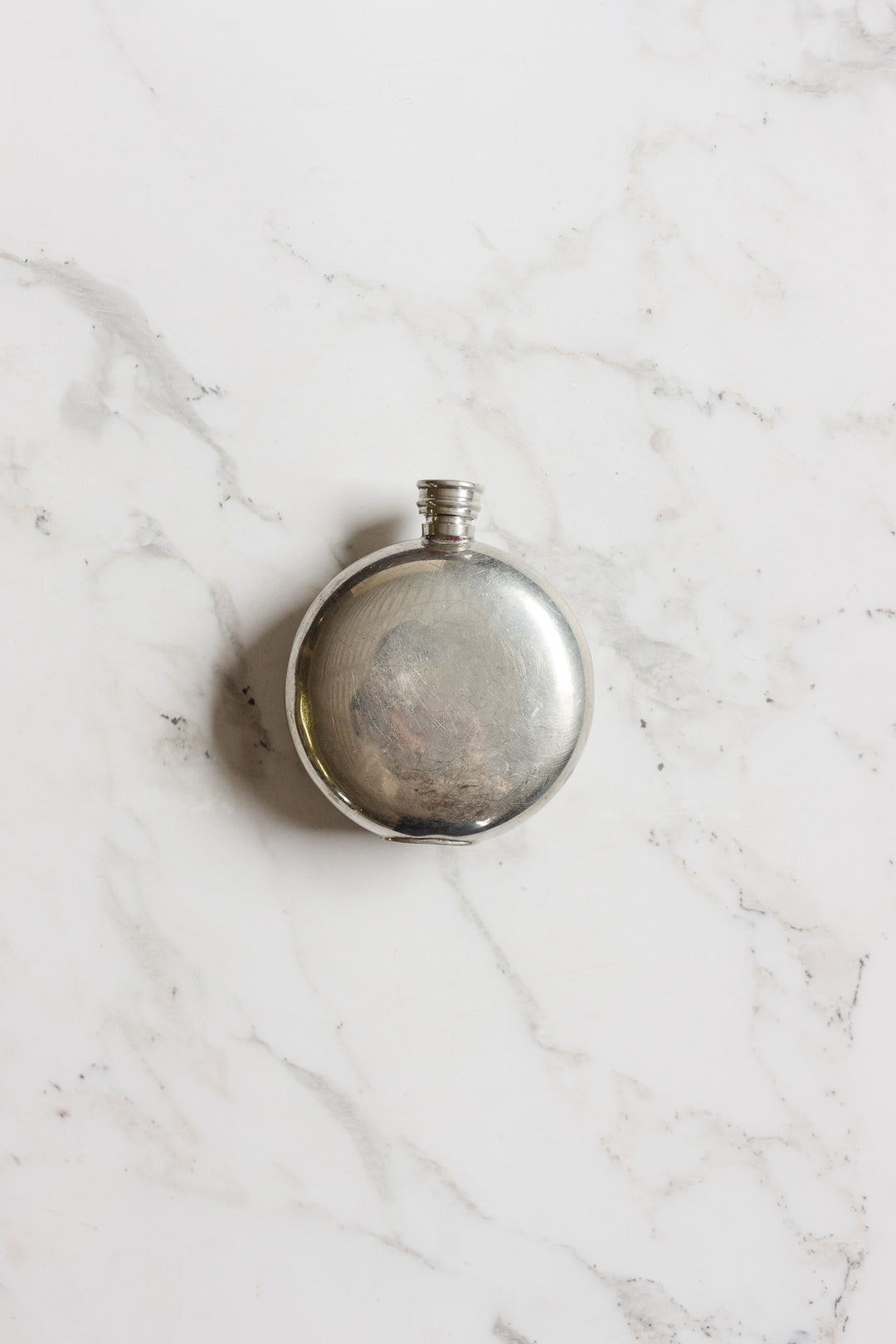 The flask is a round, silver metal vessel with a screw cap, shown centered on white marble with gray veining. Its shiny, reflective finish and compact size make this flask ideal for easy carrying.