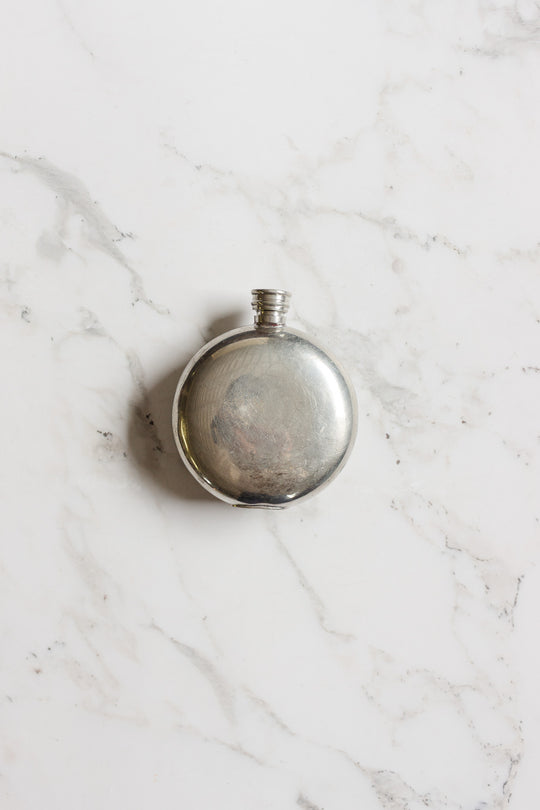 The flask is a round, silver metal vessel with a screw cap, shown centered on white marble with gray veining. Its shiny, reflective finish and compact size make this flask ideal for easy carrying.