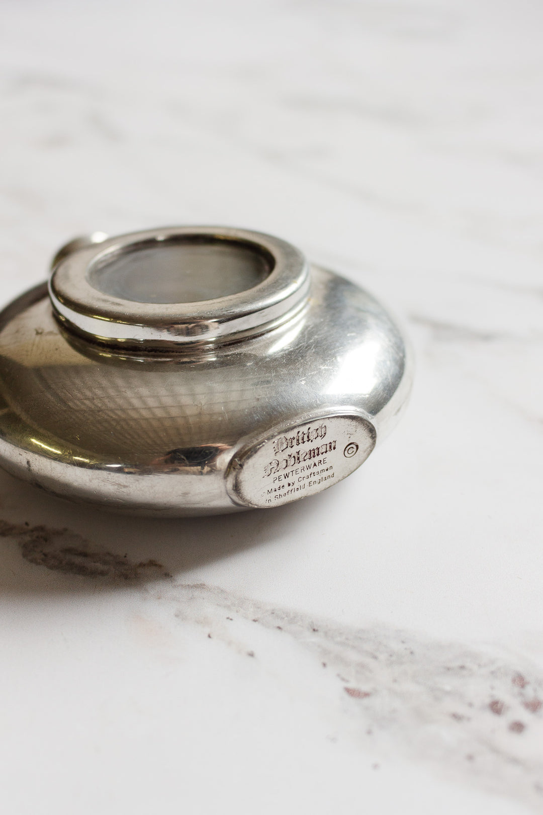 A round, silver flask with a glass window on top rests on a white marble surface. An engraved metal plaque displaying text and dimensions is visible on the side of the flask.