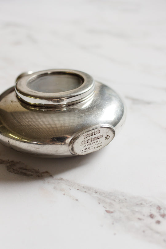 A round, silver flask with a glass window on top rests on a white marble surface. An engraved metal plaque displaying text and dimensions is visible on the side of the flask.