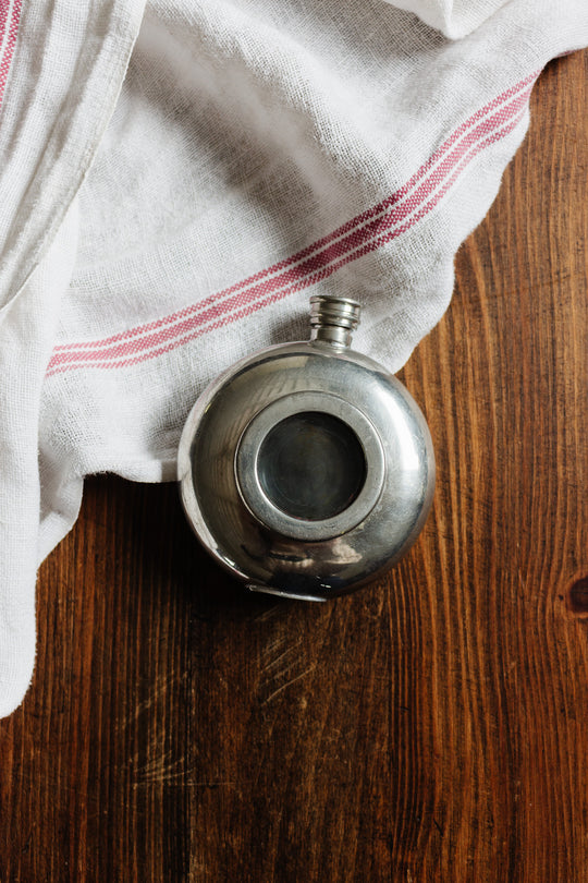 The flask, a round metal container with a screw-top lid, rests on a wooden surface beside a white cloth with red stripes; its compact size makes it ideal for travel.