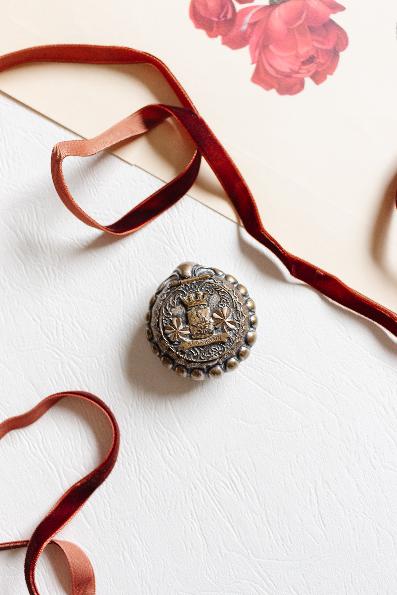 An antique French "St. Germain" pill box, featuring a round ornate metallic brooch with a crest design, is displayed on white textured fabric with red velvet ribbon and a cream paper adorned with red floral illustrations.
