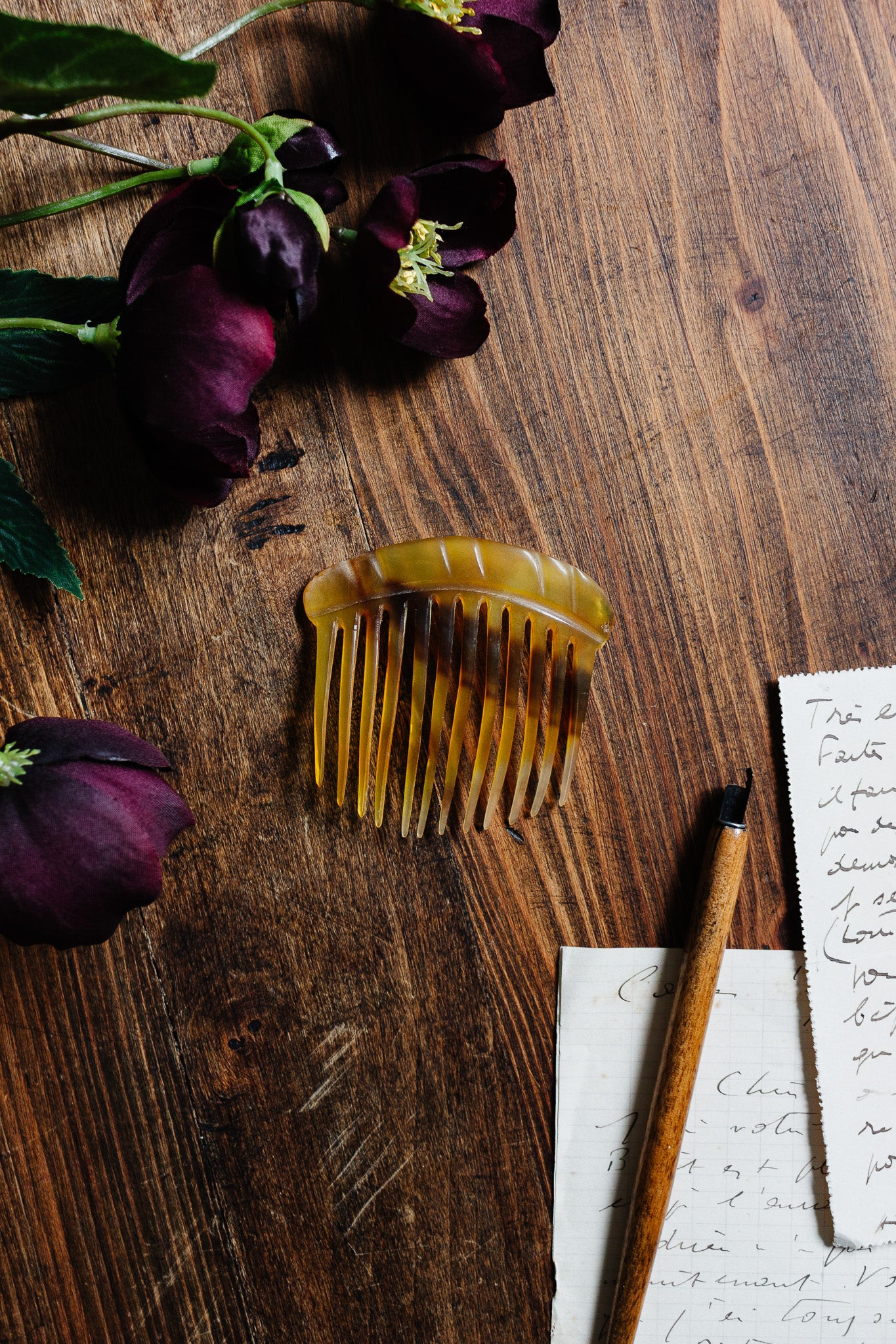 A turn of the century French horn hair comb in tortoise shell rests on a wooden table beside deep purple flowers, handwritten letters, and a wooden pen.