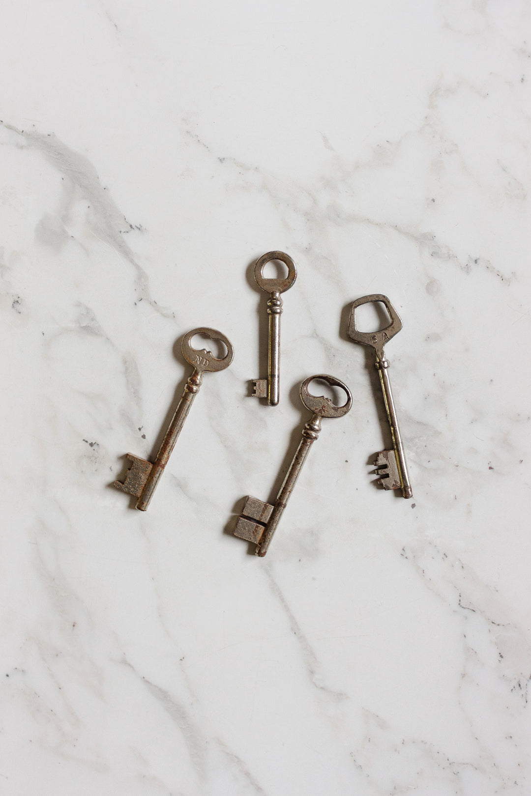 Four vintage silver keys with uniquely shaped heads are displayed on a white marble surface. Part of the keys collection, each shows signs of age and wear, highlighting their antique charm and distinctive design.