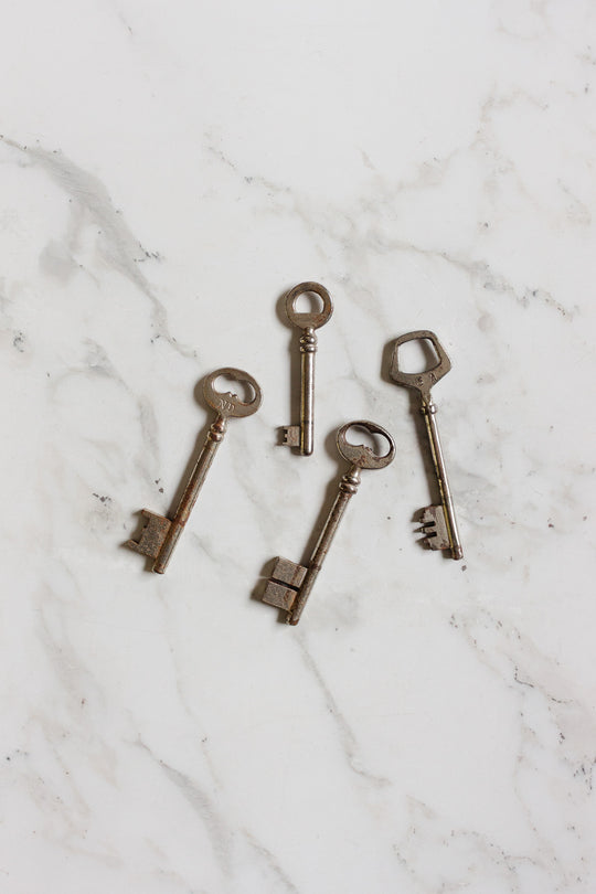 Four vintage silver keys with uniquely shaped heads are displayed on a white marble surface. Part of the keys collection, each shows signs of age and wear, highlighting their antique charm and distinctive design.