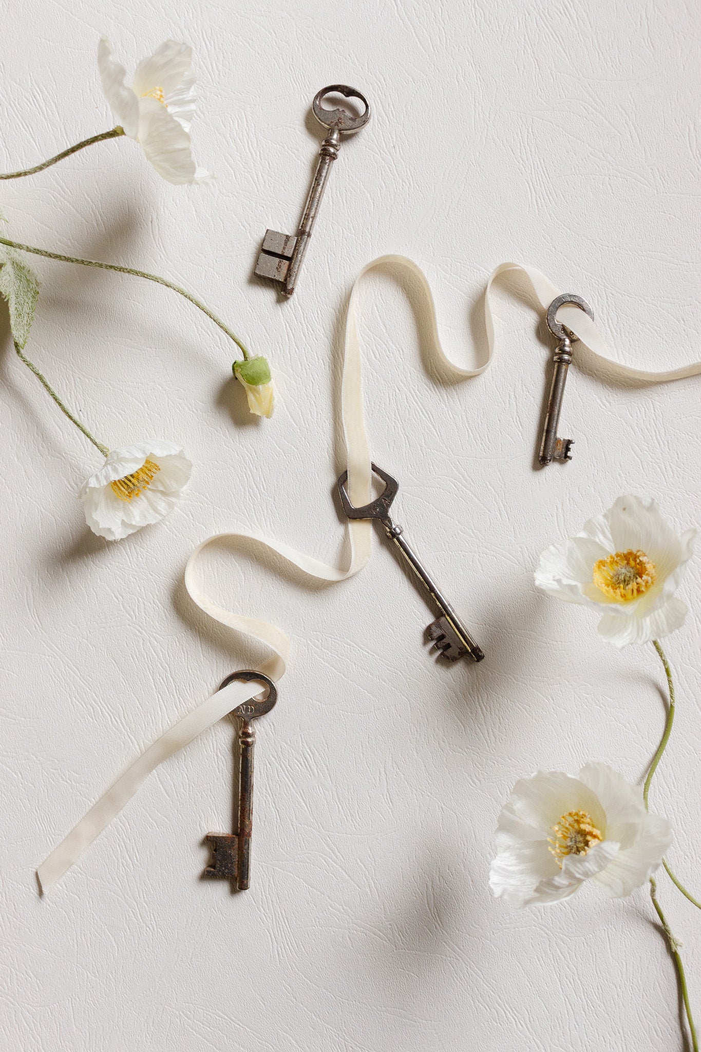 Four vintage keys are arranged with pretty white flowers on a textured surface, a cream ribbon weaving among them. The keys add charm and delicate dimension to any display.