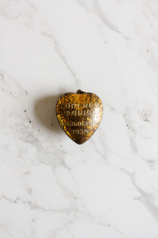 A rare antique French "coeur de marie" pendant from Boulogne, 1938, gold-colored with a vintage patina, rests on white marble—resembling a cherished religious keepsake from another era.