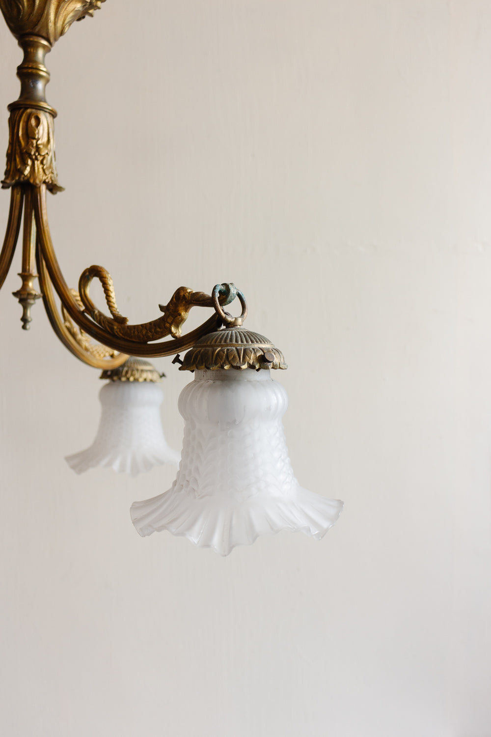 A 1920s French brass chinoiserie 3 light chandelier features ornate detailing and frosted glass ruffled-edge shades against a plain wall, with another similar shade softly blurred in the background.