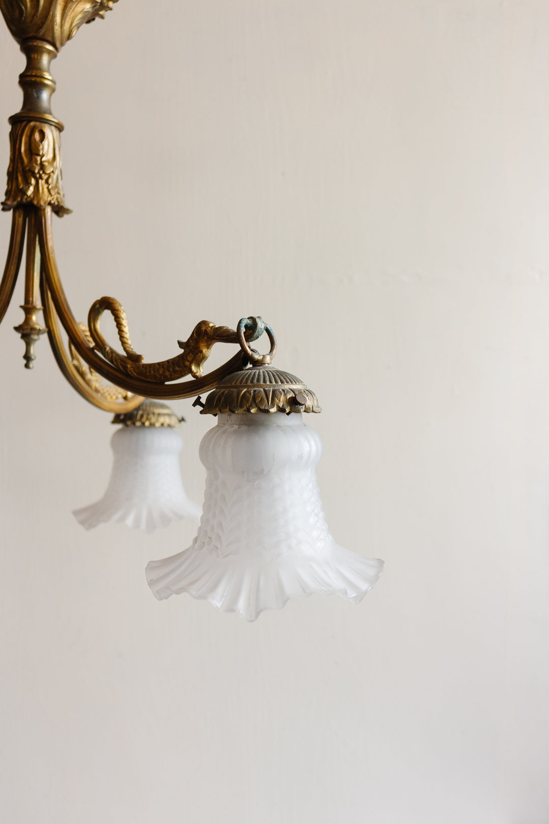 A 1920s French brass chinoiserie 3 light chandelier features ornate detailing and frosted glass ruffled-edge shades against a plain wall, with another similar shade softly blurred in the background.
