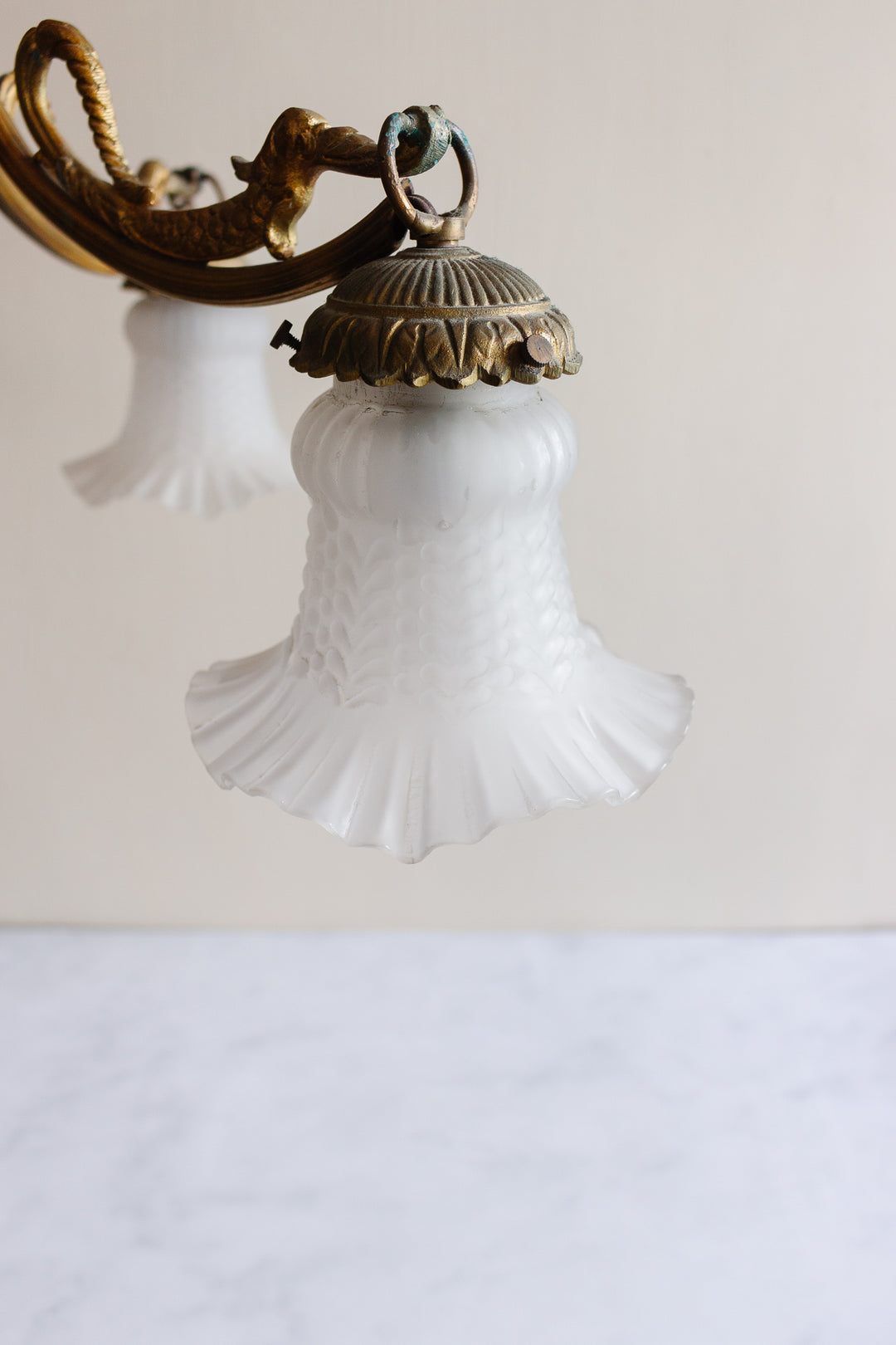 Close-up of the 1920s French brass chinoiserie 3 light chandelier, showing its ornate brass body and fluted white textured glass lampshade. A similar shade appears blurred in the background against a plain wall.