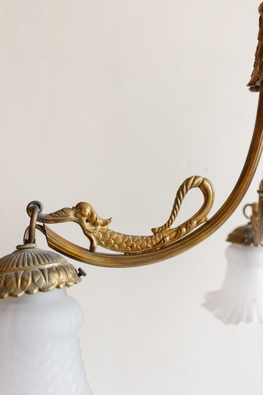 A close-up of an ornate arm from a 1920s French brass chinoiserie 3 light chandelier, shaped like a stylized fish or mythical creature, holding a frosted glass lampshade with decorative edges against a plain, light background.