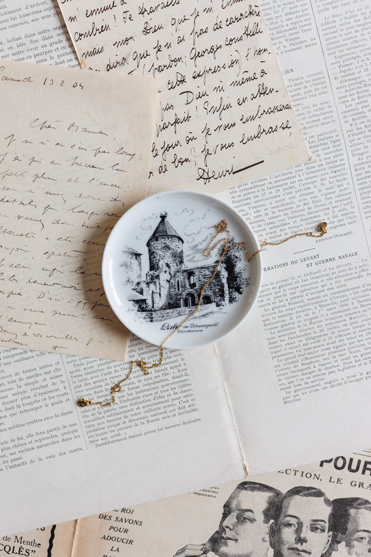 A vintage Kaiser Germany porcelain souvenir catch all with a black-and-white stone tower design sits atop handwritten letters and old newspaper, draped with a gold necklace, capturing the brocante Marseille charm.