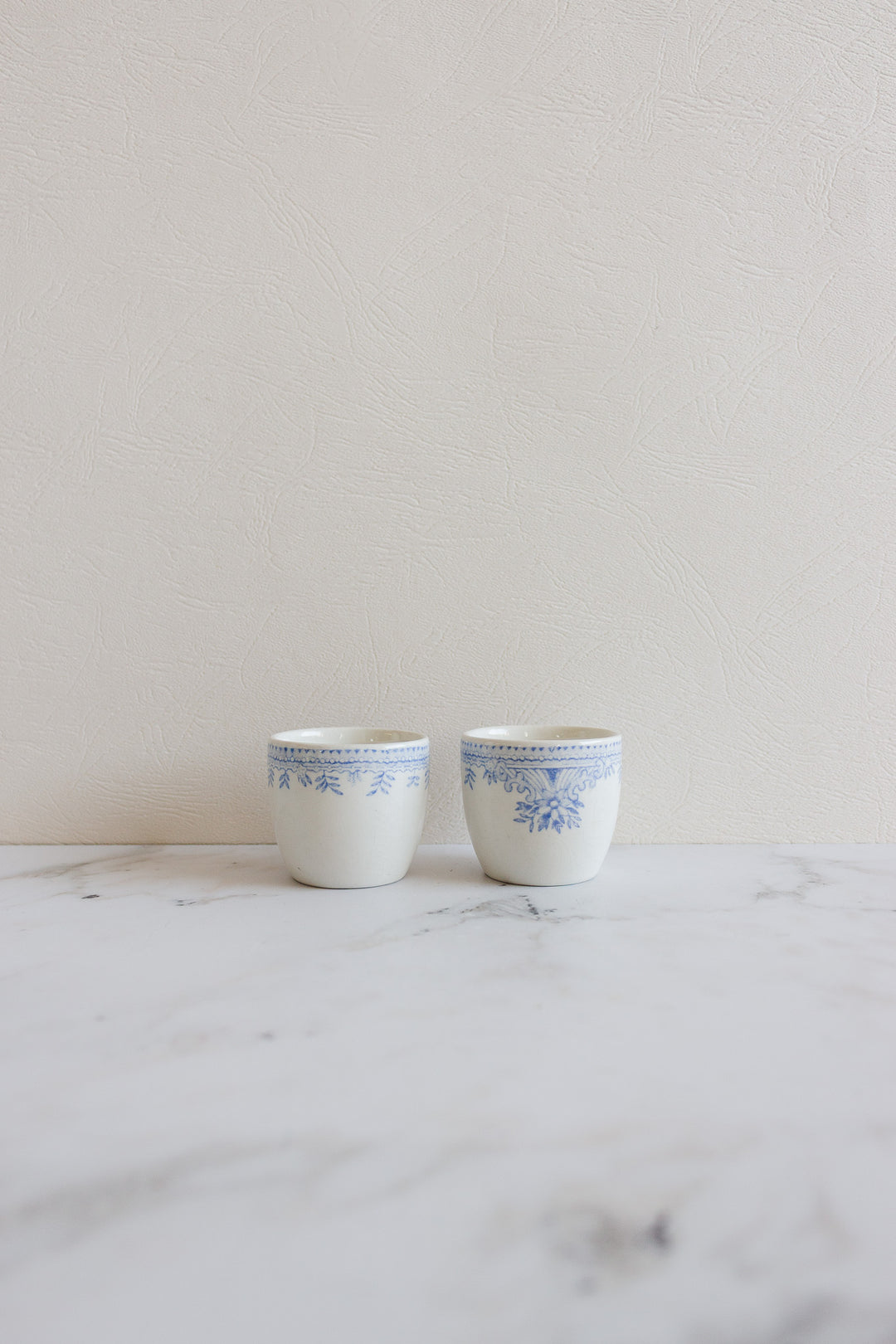 A pair of rare antique Burleigh "Asiatic Pheasants" egg cups with blue floral patterns are displayed side by side on a white marble surface against a light textured wall.