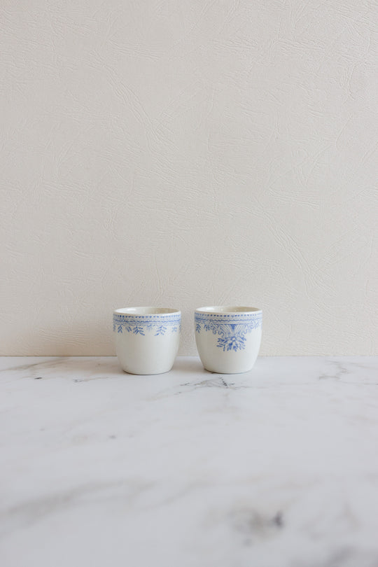 A pair of rare antique Burleigh "Asiatic Pheasants" egg cups with blue floral patterns are displayed side by side on a white marble surface against a light textured wall.