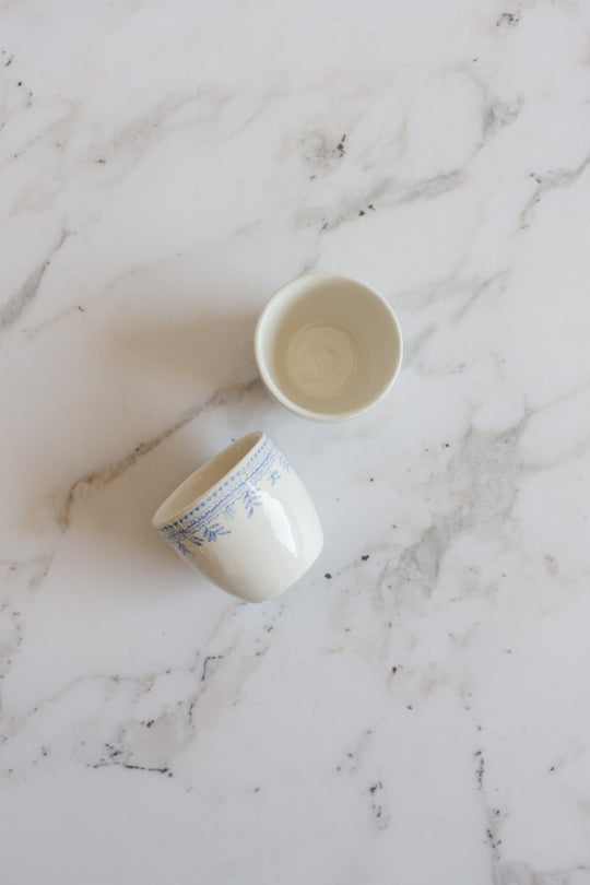 A pair of rare antique Burleigh Asiatic Pheasants egg cups sits on a white marble surface—one is plain white, the other adorned with a blue pattern near the rim inspired by the iconic Burleigh Blue Asiatic Pheasants design.