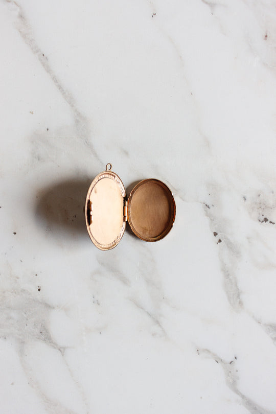An antique French onyx inlay cameo locket rests open on white marble, evoking vintage elegance. One side features a mirror, while the other contains a beige powder, suggesting it once held makeup.