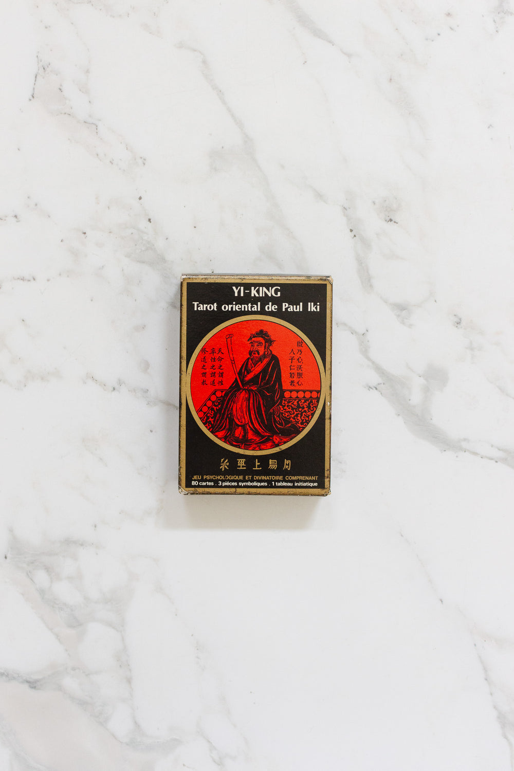 The "yi-king tarot oriental de Paul Iki cartes divinatoires" deck, featuring Asian-inspired art, is displayed in a black and red box on a white marble surface for spiritual exploration.