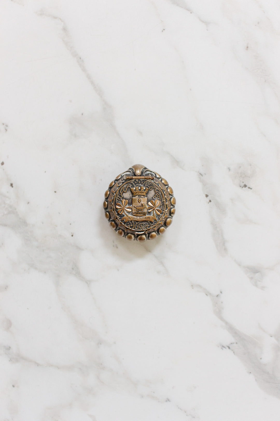 The antique French "St. Germain" pill box, a round metal object, rests elegantly on a marble surface.
