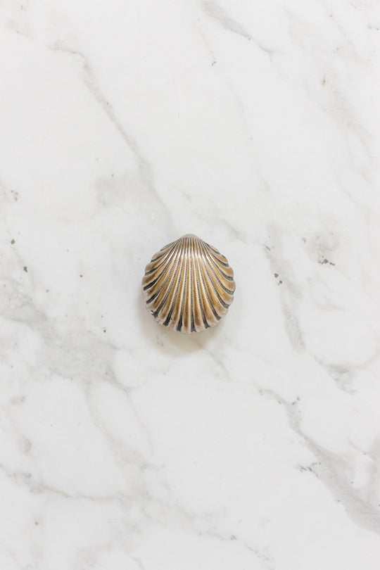 An antique French "St. Germain" pill box, featuring brown and beige ridged seashell patterns, rests on a white marble surface with light gray veining.