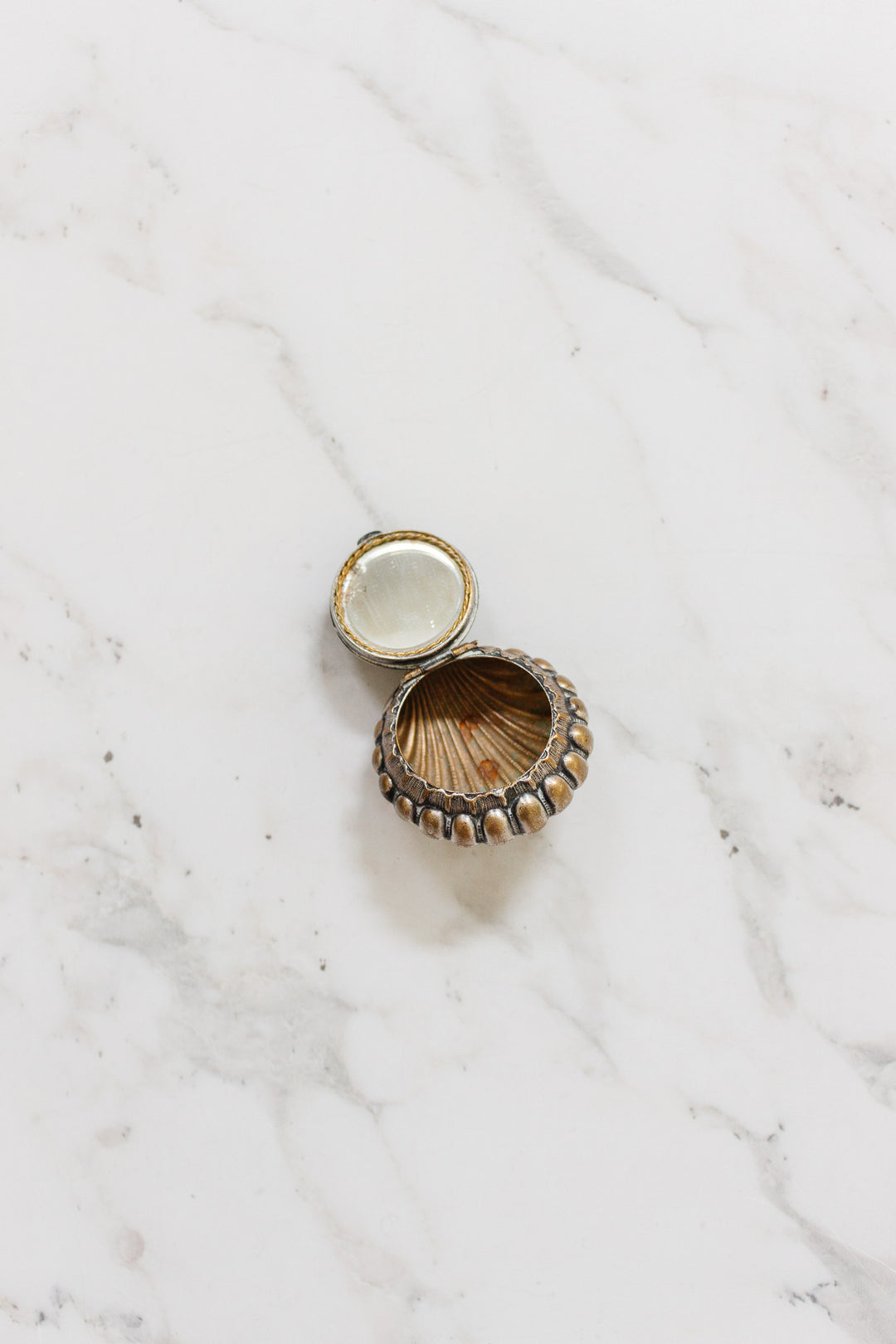 An antique French "St. Germain" pill box, small and round with a gold and bronze shell design, is displayed open on a white marble surface.