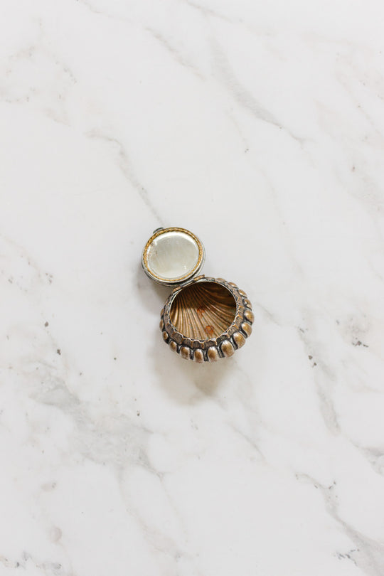 An antique French "St. Germain" pill box, small and round with a gold and bronze shell design, is displayed open on a white marble surface.