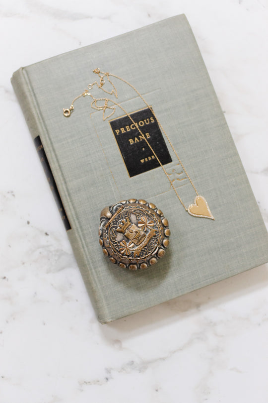 A light gray book titled Precious Bane lies on a marble surface, topped with a gold heart necklace and an antique French "St. Germain" pill box featuring a face.