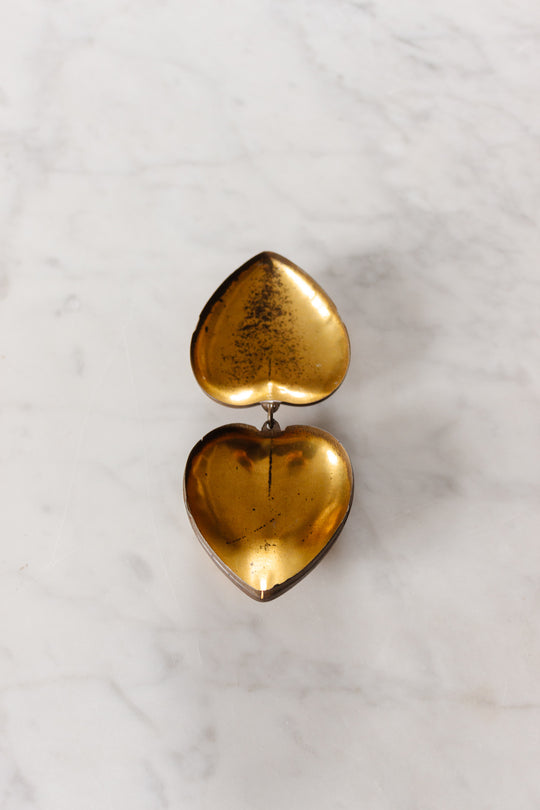 The rare antique French Coeur de Marie, Boulogne 1938 is a small, open heart-shaped gold metal box with a hinged lid and vintage charm. Its interior shows tarnishing and dark spots, sitting elegantly on a light marble surface.
