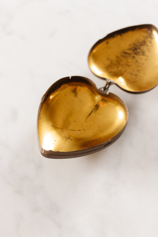 A rare antique French coeur de marie locket from Boulogne, 1938 sits open on white marble, its heart-shaped form with a golden interior showing gentle tarnish and wear, evoking vintage elegance.