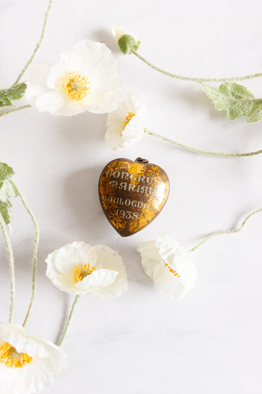 A rare antique French Coeur de Marie Boulogne 1938 heart pendant rests on a white surface, surrounded by delicate white flowers with yellow centers.