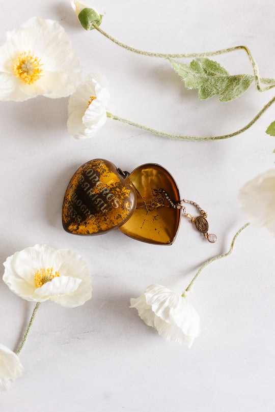 A rare antique French Coeur de Marie locket from Boulogne, 1938, lies open on a white surface with a delicate gold necklace inside. White poppy flowers surround this timeless religious keepsake.