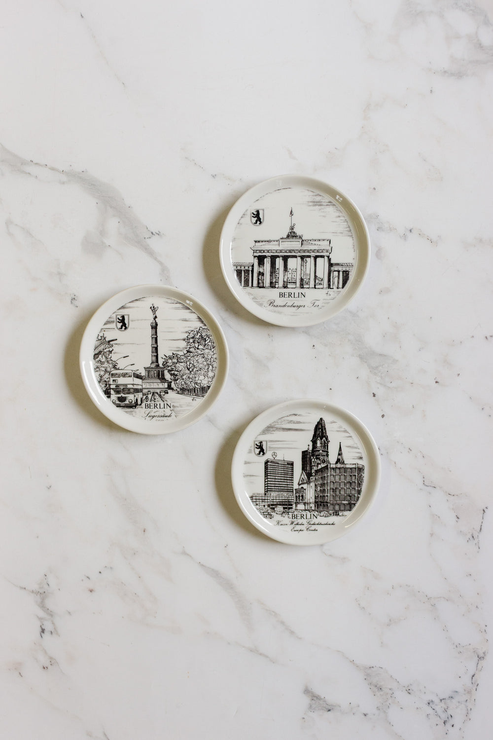 Three german coasters, each with black-and-white drawings of iconic Berlin landmarks and minimal text, are displayed on a white marble surface. Each coaster features a unique design and size.