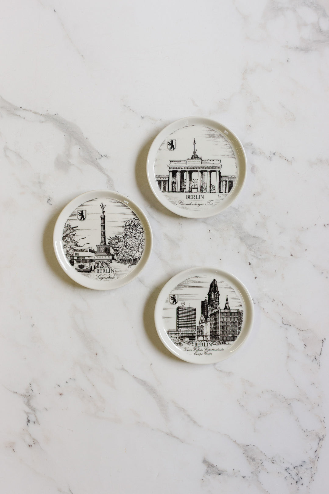 Three german coasters, each with black-and-white drawings of iconic Berlin landmarks and minimal text, are displayed on a white marble surface. Each coaster features a unique design and size.
