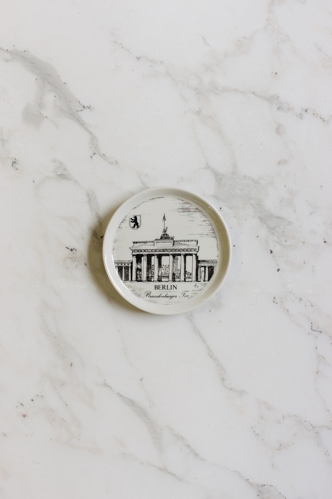 A german coaster, small and round, features a black-and-white illustration of Berlin’s Brandenburg Gate with "Berlin" written below, displayed on a white marble surface.